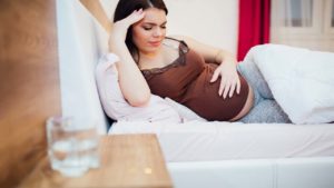 woman with headache lying in bed with hand over pregnant stomach